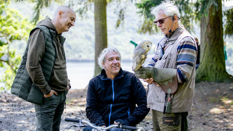 Drei Personen im Kreis. Eine Person hält eine Eule auf der Hand. Eine Person sitzt in einem Rollstuhl.