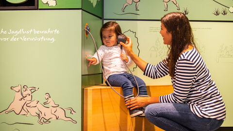 Kind sitzt in Kellerwalduhr auf Bank mit Lautsprecher am Ohr