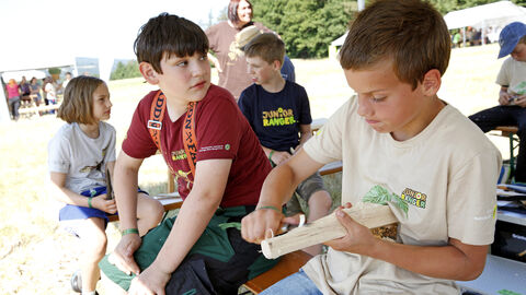 Mehrere Kinder sitzen zusammen, ein Kind schnitzt an einem Stück Holz mit einem Messer