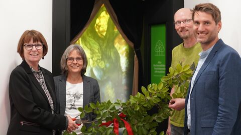 Vier Person (links zwei Frauen, rechts zwei Männer) stehen vor einer Tür. Zwischen ihnen ist ein rotes Band mit Ästen dazwischen.