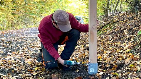 Rettungskette wird sichergestellt: Aufgrund der längeren Bauphase am Oberbecken Waldeck I installiert die Nationalparkverwaltung ein festes Läufersystem entlang der Umleitungen im Schutzgebiet. Die Holzpfosten enthalten neben dem Wegesymbol auch eine grüne Plakette mit dem Kürzel „NLP“ und einer 4-stelligen Nummer, die der Rettungsstelle im Notfall Auskunft über den Standort im Schutzgebiet gibt. Mitarbeiter schreibt Holzpfosten im Wald in den Boden