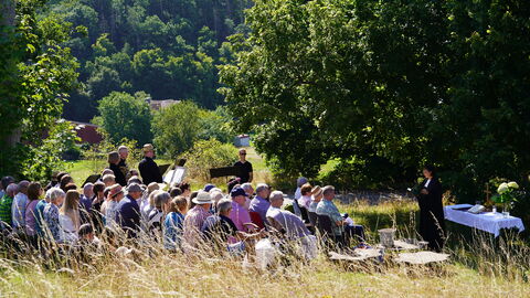 Den Gottesdienst an der Linde auf der Koppe hat der Posaunenchor Die Neuen Alten aus Frankenau musikalisch begleitet. Menschen sitzen auf Bänken auf einer sonnigen Wiese neben einem großen Baum, während die Pfarrerin predigt und der Posaunenchor auf seinen Einsatz wartet.