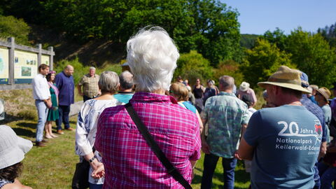 Start der Wanderung und Begrüßung an der Koppe Eine Gruppe Menschen in Wanderkleidung steht im Halbkreis um eine Wandertafel in der Sonne