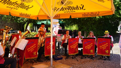 Unter einem gelben Sonnenschirm sitzen Blechbläser und spielen ihr Instrument. Auf einem roten Banner, das vor jedem in der ersten Reihe steht, steht der Name der Kapelle