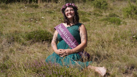 Heideblütenprinzessin Saija I ist die insgesamt vierte Altenlotheimer Heideblütenprinzessin Eine junge Frau mit dunklen, schulterlangen Haaren in einem perolfarbenen Kleid und einem Haarkranz aus Heidekraut im Haar sitzt auf einer Wiese, auf der einzelne Heideblütenbüschel blühen