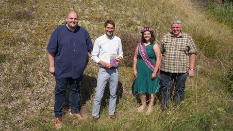 Die Heideblütenprinzessin steht mit drei Männern an einem bewachsenen Hügel in der Sonne, alle lächeln in die Kamera