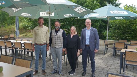 Drei Männer und eine Frau stehen nebeneinander unter aufgespannten Sonnenschirmen auf einer Terrase mit Möbeln