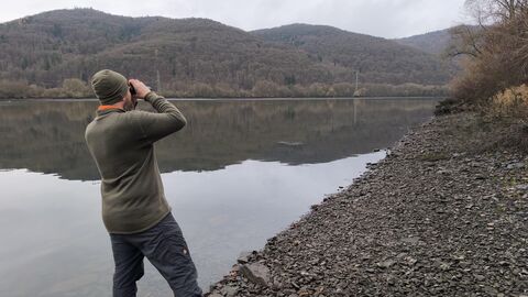in einer grauen Landschaft am steinigen Seeufer steht ein Mann mit Rücken zur Kamera und blickt mit einem Fernglas über den See. Im Hintergrund erkennt man die bewaldeten Hügel des Nationalparks - in dieser Jahreszeit ohne Blätter an den Bäumen.
