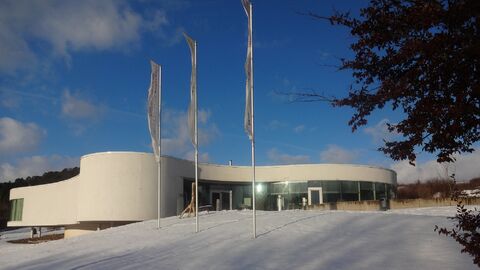 Das futuristische NationalparkZentrum in verschneiter Landschaft vor blauem Himmel. An den 3 Fahnenmasten davor flattern Fahnen im Wind.
