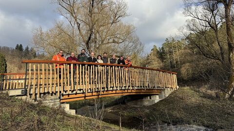 Auf einer Brücke über einen Bach stehen 9 Personen und lächlen in die Kamera. Die Bäume im Hintergrund haben noch keine Blätter und die tiefstehende Sonne scheint allen ins Gesicht. Das Brückengeländer besteht aus senkrecht angebrachten Holzstämmchen ohen Rinde