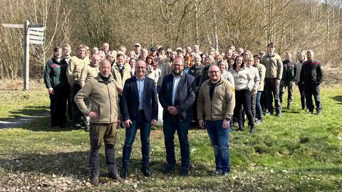4 Männer stehen auf einer Wiese im Vordergrund in der Sonne, im Hintergrund stehen circa 50 Menschen als Gruppe in Imagekleidung: die Mitarbeitenden des Nationalparks.