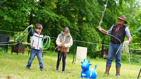 Ein Cowbow und 2 kleine Kinder haben ein Lasso in der Hand und im Vordergrund ist eine kniehohe blaue Gummikuh zu sehen