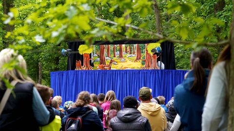 Vor grünen Laubbäumen steht eine Bühne mit Figuren auf der Bühne. Die Zuschauer sitzen davor und sind von hinten zu sehen.