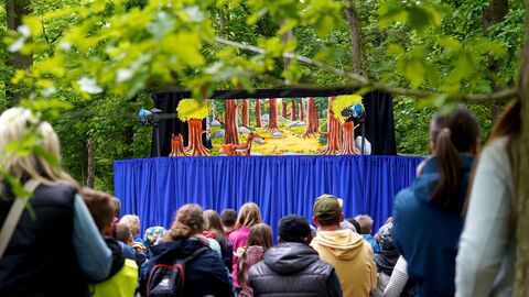 Vor grünen Laubbäumen steht eine Bühne mit Figuren auf der Bühne. Die Zuschauer sitzen davor und sind von hinten zu sehen.