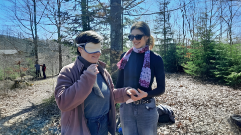 Zwei junge Frauen stehen in einem sonnigen Waldstück. Eine hat die Augen verbunden, die andere führt sie an der Hand. Beide lächeln.