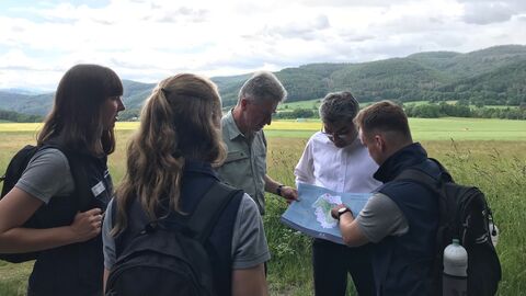 5 Personen stehen um eine Landkarte und blicken darauf. Im Hintergrund erkennt man die bewaldeten Hügel des Nationalparks.