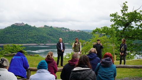 Auf einer Wiese stehen 4 Personen. Zwei Männer, eine Gebärdendolmetscherin, die Gesten mit der Hand formt und die Falknerin. Im Hintergrund der Edersee und Schloss Waldeck.