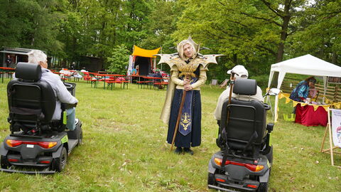 Von hinten sieht man eine ältere Dame und einen älteren Herren auf einem E-Scooter sitzen. Sie sind im Gespräch mit einer blonden Dame in einem fantasievollen Kostüm.
