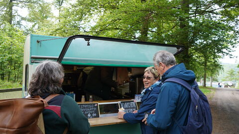 An der Verkaufsluke eines Mini-Carava-Anhängers in türkis stehen 3 Personen und warten auf ihren Kaffee.