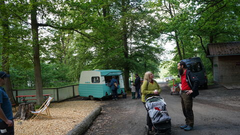 Szene im WildtierPark: Im Vordergrund unterhalten sich 2 Personen. Im Hintergrund grüne Bäume und der türkisfarbene Cafe-Caravan.