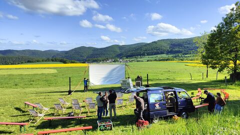 Im Vordergrund eine grüne Wiese, die in der Sonne liegt. Im Hintergrund die bewaldeten Hügel des Nationalparks. Auf der Wiese sind eine Kinoleinwand und Sitzbänke und -stühle aufgestellt.