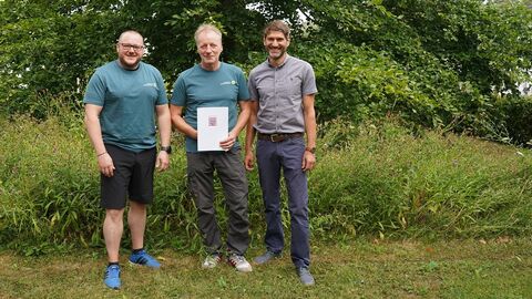 3 Männer in sommerlichen Outdoor-Sachen stehen vor einem grünem Baum und hohem Gras. Der mittlere hält eine Urkunde vor sich und alle lächeln in die Kamera.