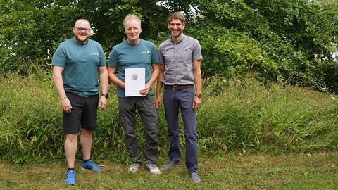 3 Männer in sommerlichen Outdoor-Sachen stehen vor einem grünem Baum und hohem Gras. Der mittlere hält eine Urkunde vor sich und alle lächeln in die Kamera.