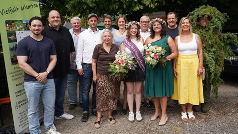 Zweireihiges Gruppenfoto: vorne stehen 4 Frauen in sommerlichen Kleidern, die beiden in der Mitte tragen einen Blütenkranz mit Heide und je einen Blumenstrauß, eine trägt eine Schärpe. In der hinteren Reihe stehen bis auf eine Frau nur Männer, einer davon ist vor lauter Wacholderbüschen gar nicht zu erkennen, nur das Gesicht schaut raus. Alle lächeln in die Kamera.