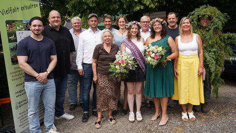 Zweireihiges Gruppenfoto: vorne stehen 4 Frauen in sommerlichen Kleidern, die beiden in der Mitte tragen einen Blütenkranz mit Heide und je einen Blumenstrauß, eine trägt eine Schärpe. In der hinteren Reihe stehen bis auf eine Frau nur Männer, einer davon ist vor lauter Wacholderbüschen gar nicht zu erkennen, nur das Gesicht schaut raus. Alle lächeln in die Kamera.