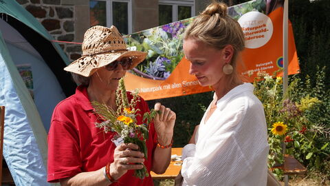 Eine Frau in rotem T-Shjort und mit Strohhut hat ein Büschel aus blühenden Kräutern in der Hand, eine zweite Frau guckt interessiert.