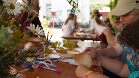 An einem Tisch sitzen ein Mädchen und eine Frau und haben buntes Papier in der Hand. In den Vordergrund ragt ein Strauß Wiesenblumen.