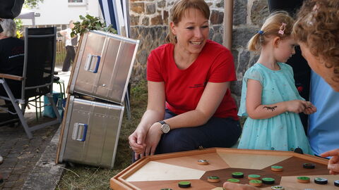 Zwei Frauen sitzen und ein Mädchen stehen an einem mehreckigen Holzbrett mit Spielsteinen.