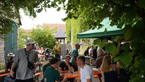Blick in einen mit Büschen umrandeten Teil des Festgebiets. Am Rand stehen Tische, an denen Leute sitzen, in der Mitte ein kleiner nachgebauter Turm der Quernstkapelle