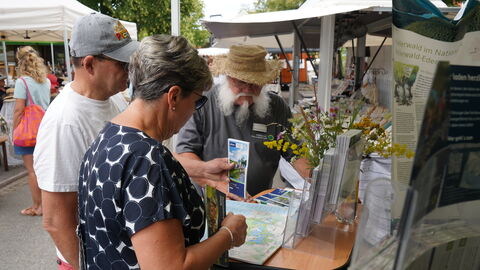 Ein Paar steht an einem Infostand und hält einen Flyer in der Hand. Ein weißbärtiger Mann spricht mit ihnen