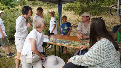 Menschen sitzen an einer Bierzelttischgarnitour unter einem Sonnenschutz in hügeliger Heidelandschaft