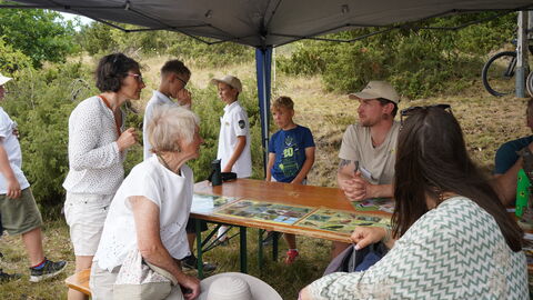 Menschen sitzen an einer Bierzelttischgarnitour unter einem Sonnenschutz in hügeliger Heidelandschaft
