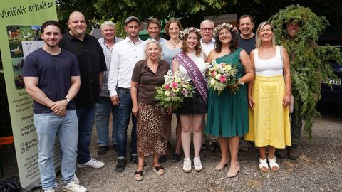 Zweireihiges Gruppenfoto: vorne stehen 4 Frauen in sommerlichen Kleidern, die beiden in der Mitte tragen einen Blütenkranz mit Heide und je einen Blumenstrauß, eine trägt eine Schärpe. In der hinteren Reihe stehen bis auf eine Frau nur Männer, einer davon ist vor lauter Wacholderbüschen gar nicht zu erkennen, nur das Gesicht schaut raus. Alle lächeln in die Kamera.