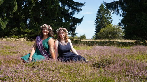 Zwei junge Frauen sitzen in blühendem Heidekraut: die links sitzenden hat dunkelbraune Haare mit leichten Wellen und ein petrolfarbenes Kleid, die rechts dunkelblonde Haare mit leichten Wellen und ein dunkelblaues Kleid. Die links trägt eine beerenfarbene Schärpe, beide haben Blumenkränze auf und lächeln in die Kamera.