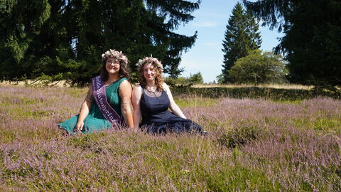 Zwei junge Frauen sitzen in blühendem Heidekraut: links sie hat dunkelbraune Haare mit leichten Wellen und ein petrolfarbenes Kleid, die rechts dunkelblonde Haare mit leichten Wellen und ein dunkelblaues Kleid. Die links trägt eine beerenfarbene Schärpe und beide haben Blumenkränze auf.