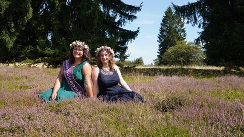 Zwei junge Frauen sitzen in blühendem Heidekraut: links sie hat dunkelbraune Haare mit leichten Wellen und ein petrolfarbenes Kleid, die rechts dunkelblonde Haare mit leichten Wellen und ein dunkelblaues Kleid. Die links trägt eine beerenfarbene Schärpe und beide haben Blumenkränze auf.