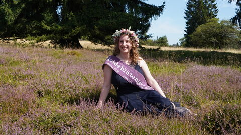 eine junge Frau sitzt in blühendem Heidekraut und lächelt in die Kamera. Sie trägt ein dunkelblaues Kleid, eine heidekrautfarbene Schärpe und einen Blumenkranz im Haar.