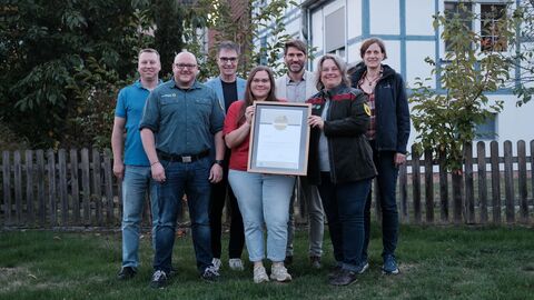 Gruppenfoto mit 4 Männern und 3 Frauen. Alle lächeln in die Kamera, die beiden Frauen in der Mitte halten einen großen Bilderrahmen mit einer Urkunde.