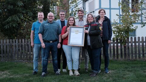 Gruppenfoto mit 4 Männern und 3 Frauen. Alle lächeln in die Kamera, die beiden Frauen in der Mitte halten einen großen Bilderrahmen mit einer Urkunde.